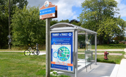 Lansing River Trail access sign in front of a CATA Transit to Trails bus shelter with bike and trail in the background