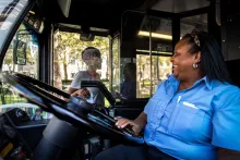 A smiling bus driver in a blue uniform interacts with a passenger through a clear protective barrier.